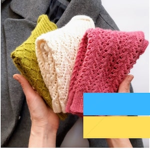 May include: Three knitted scarves in different colors: a mustard yellow, a cream white, and a pink. The scarves are folded and held in a person's hand.