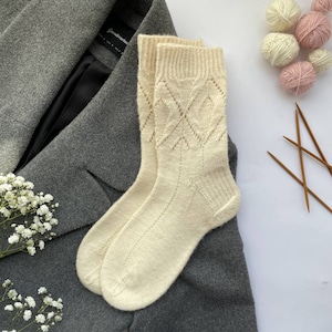 Puede incluir: Un par de calcetines de punto de color blanco roto con un patrón de encaje en forma de diamante. Los calcetines están sobre una superficie de tela gris.