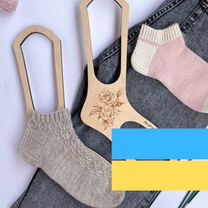 May include: Two wooden sock blockers with a floral design. One sock blocker is holding a beige sock and the other is holding a pink and white sock. The sock blockers are on top of a pair of blue jeans.