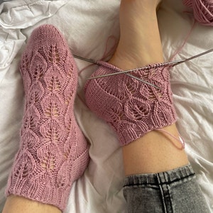 Könnte beinhalten: Ein Paar rosa gestrickte Socken mit einem Spitzenmuster. Die Socken werden mit Stricknadeln gearbeitet.