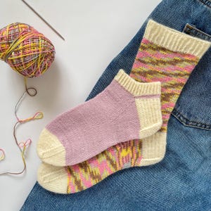 May include: Two pairs of hand-knitted socks, one pair is pink and white, the other is a multi-colored pattern of pink, yellow, brown, and white. The socks are laying on a pair of blue jeans.