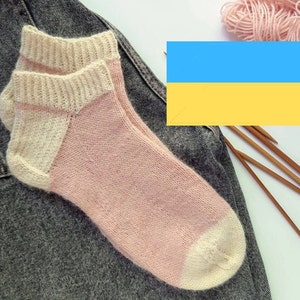 Può includere: Un paio di calzini di lana rosa e bianchi con tallone e punta a contrasto. I calzini sono appoggiati su un pezzo di tessuto di jeans. Sullo sfondo c'è un gomitolo di lana rosa e ferri da maglia.