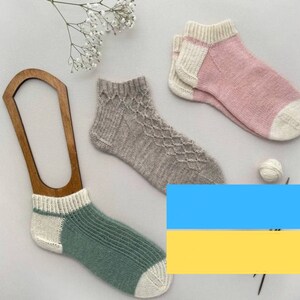 May include: A collection of handmade socks in various colors, including pink, gray, and green, displayed with a wooden sock form. The socks feature different knit patterns and are made with soft, textured yarn. A small ball of yarn and sprigs of white flowers are also present.