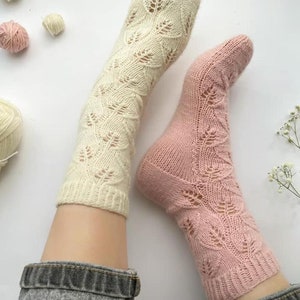 May include: A pair of knitted socks with a delicate leaf pattern. One sock is cream coloured and the other is pink. The socks are shown on a person's legs, with the tops of the socks folded down.
