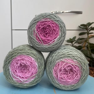 May include: Three round balls of yarn stacked in a pyramid formation. Each ball features a gradient colour pattern, transitioning from a soft sage green on the outer layers to a vibrant magenta pink at the centre, with thin white accents.