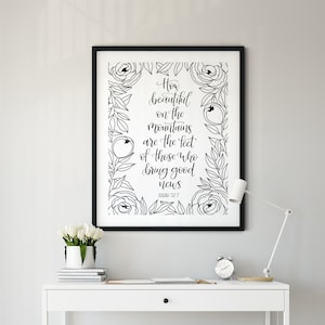 Peut inclure: Une impression encadrée avec le texte "How beautiful on the mountains are the feet of those who bring good news" dans une écriture élégante, entourée d'illustrations florales. L'impression est au-dessus d'un bureau blanc avec une chaise, une lampe et un vase de fleurs blanches.