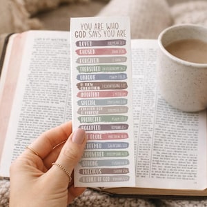 Könnte beinhalten: Ein Lesezeichen mit dem Text "YOU ARE WHO GOD SAYS YOU ARE" und verschiedenen Affirmationen. Das Lesezeichen wird über einer offenen Bibel neben einer gesprenkelten Tasse mit einem Getränk gehalten. Der Hintergrund ist eine weiche, neutrale Farbe.