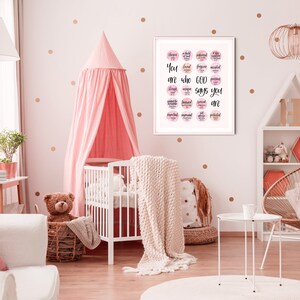May include: A white crib with a pink canopy and a white blanket. A framed print with pink and white watercolor circles and the text "You are who God says you are" hangs on the wall. The wall is decorated with gold polka dots.