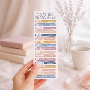 Könnte beinhalten: Ein weißes Lesezeichen mit dem Text "YOU ARE WHO GOD SAYS YOU ARE" und einer Liste von Affirmationen in verschiedenen Farben. Das Lesezeichen wird von einer Hand gehalten, mit Büchern und einer Tasse im Hintergrund.
