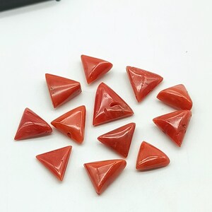 Puede incluir: Una colección de gemas triangulares de coral pulido. Cada gema es de un tono rojo vibrante, con superficies lisas y reflectantes. Las gemas están dispuestas sobre una superficie blanca, mostrando su forma y color.