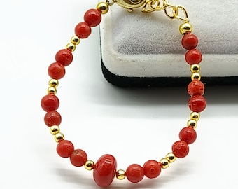 Puede incluir: Una pulsera de cuentas de coral rojo con detalles dorados. La pulsera presenta pequeñas cuentas rojas redondas intercaladas con cuentas doradas. Una cuenta roja más grande, de forma ovalada, está centrada. El cierre y la cadena son dorados.