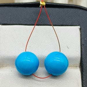 Pode incluir: Um par de brincos esféricos azuis turquesa brilhantes. Os brincos são enfiados em um fio fino, marrom-avermelhado, que se conecta no topo com uma pequena conta dourada. Os brincos são exibidos em um fundo neutro.