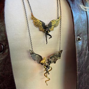 May include: Two necklaces with detailed pendant designs. The top necklace features a figure with gold and black wings. The bottom necklace has a similar design, with a figure and a golden serpent. Both necklaces have dark chains.