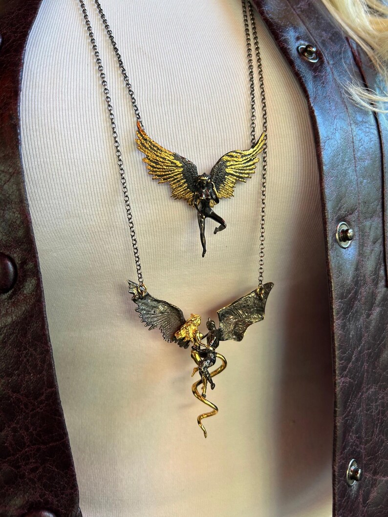 May include: Two necklaces with detailed pendant designs. The top necklace features a figure with gold and black wings. The bottom necklace has a similar design, with a figure and a golden serpent. Both necklaces have dark chains.