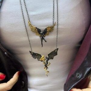 May include: Two layered necklaces with angel pendants. The top necklace features a black angel with gold wings. The bottom necklace has a black angel with gold accents and a gold serpent. The chains are silver-toned.