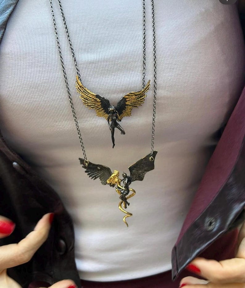 May include: Two layered necklaces with angel pendants. The top necklace features a black angel with gold wings. The bottom necklace has a black angel with gold accents and a gold serpent. The chains are silver-toned.