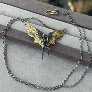 May include: A silver-toned necklace featuring a pendant of a winged figure with gold-colored wings. The figure is in a dynamic pose, and the necklace has a delicate chain with a lobster clasp.