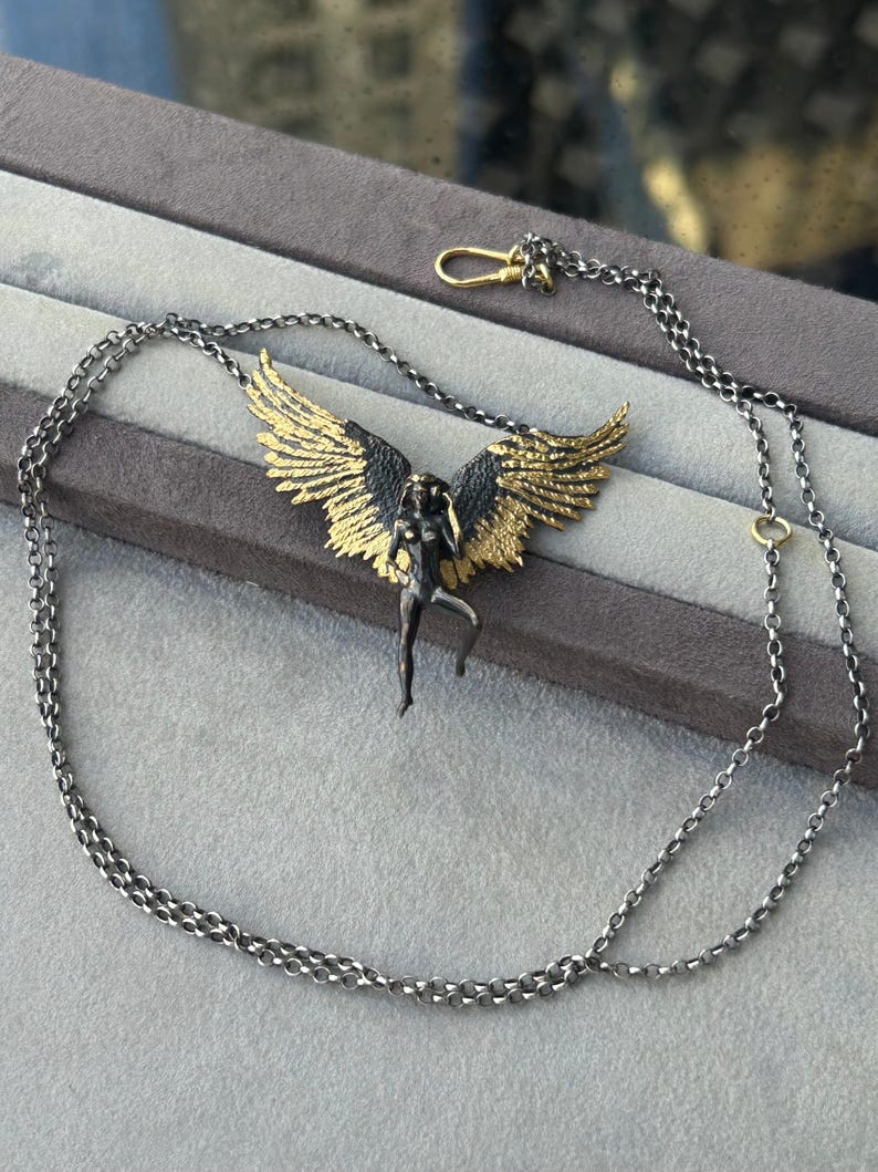 May include: A silver-toned necklace featuring a pendant of a winged figure with gold-colored wings. The figure is in a dynamic pose, and the necklace has a delicate chain with a lobster clasp.
