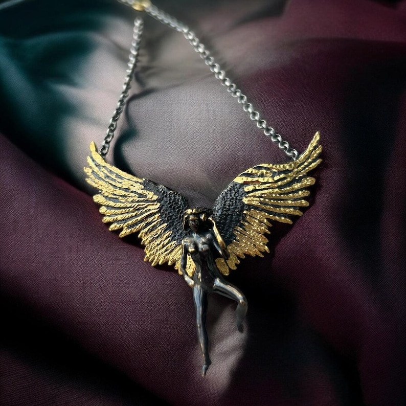 May include: A silver-toned necklace featuring a stylized figure with outstretched wings. The wings are detailed with gold and black accents, and the figure is posed in a graceful stance. The necklace hangs against a dark, textured background.
