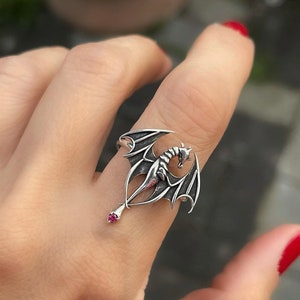 May include: A silver dragon ring with a pink gemstone. The dragon is in a flying pose with its wings spread out. The ring is worn on a finger.