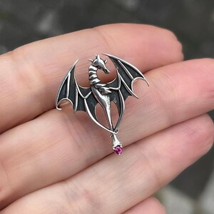 May include: A silver dragon pendant with outstretched wings and a pink gemstone at the end of its tail.
