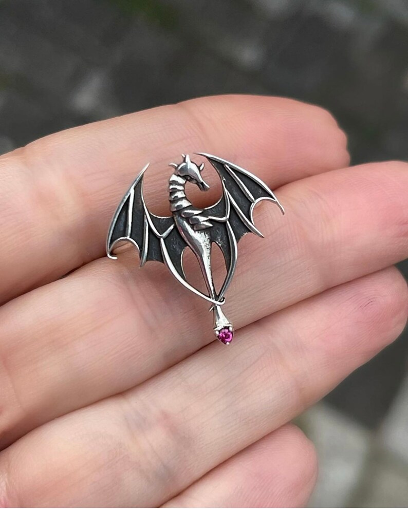 May include: A silver dragon pendant with outstretched wings and a pink gemstone at the end of its tail.