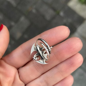 May include: A silver ring featuring a dragon design. The dragon is curled around a silver band, and the ring is open on one side.