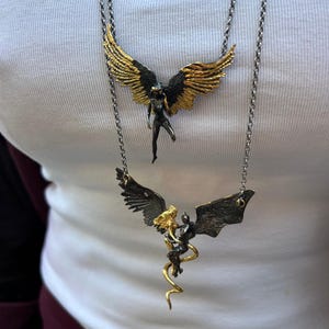 May include: Two necklaces with angel and demon pendants. The top necklace features a gold-winged angel pendant with a black body. The bottom necklace has a gold and black demon pendant with a gold serpentine design.