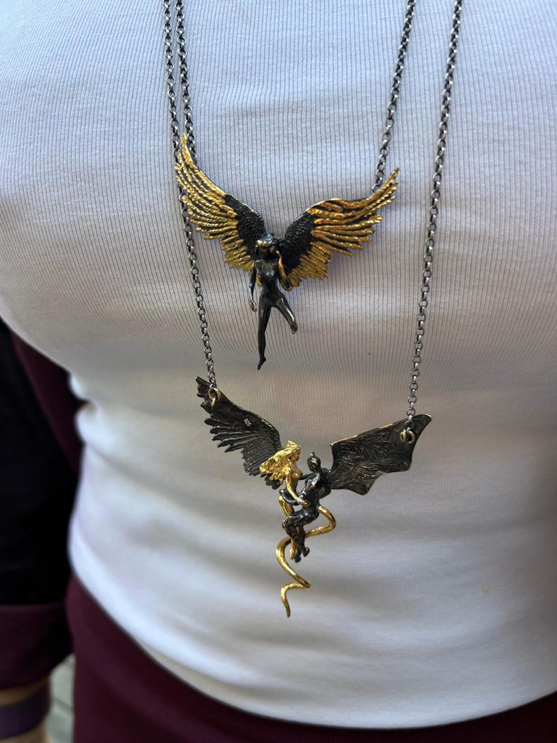 May include: Two necklaces with angel and demon pendants. The top necklace features a gold-winged angel pendant with a black body. The bottom necklace has a gold and black demon pendant with a gold serpentine design.