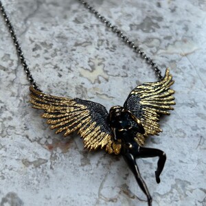 May include: A pendant necklace featuring a bronze-colored figure with outstretched wings. The wings are detailed with gold accents, and the figure is suspended from a delicate chain. The necklace has a vintage or antique aesthetic.