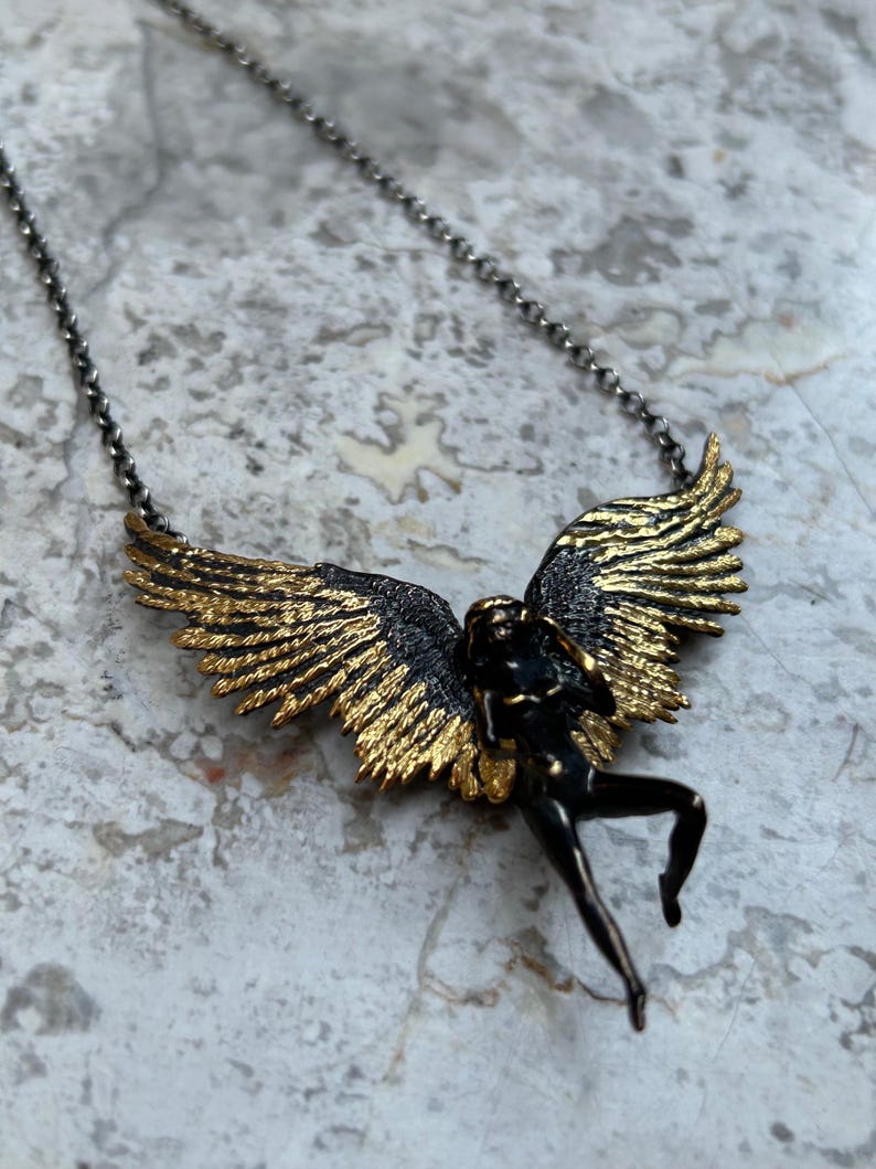 May include: A pendant necklace featuring a bronze-colored figure with outstretched wings. The wings are detailed with gold accents, and the figure is suspended from a delicate chain. The necklace has a vintage or antique aesthetic.