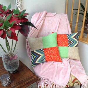 May include: Two patchwork pillows with a green, orange, and blue geometric pattern sit on a pink throw blanket on a chair. The chair is in front of a white wall and a wooden railing.
