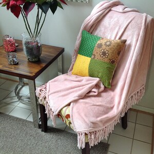 May include: A pink throw blanket with fringe is draped over a chair, with a patchwork pillow in shades of green, yellow, and brown. The chair is set in a room with a side table and a poinsettia plant in a glass vase.