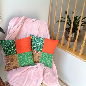 May include: Two square throw pillows with patchwork designs. One pillow features green polka dots and orange fabric, while the other has green floral and brown floral patterns. The pillows rest on a pink blanket draped over a chair.