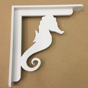 Puede incluir: Soporte de estante de madera blanco con un diseño de caballito de mar. El soporte tiene forma de caballito de mar con una cola enroscada.