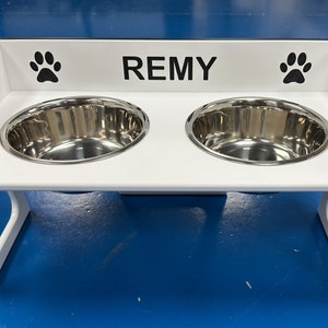 Könnte beinhalten: Ein weißer erhöhter Hundenapfständer mit zwei Edelstahlnäpfen. Der Ständer hat einen Ausschnitt für einfachen Zugang und der Name "REMY" ist mit Pfotenabdrücken auf beiden Seiten auf der Vorderseite aufgedruckt.
