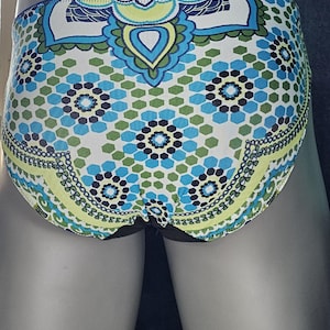 May include: A pair of patterned underwear with a colorful design. The fabric features a repeating pattern of blue, green, and white geometric shapes and floral motifs. The waistband and leg openings are trimmed with a black fabric.