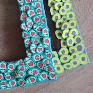 May include: A picture frame decorated with green and yellow avocado-shaped charms. The avocado charms have a cute face with a heart and eyes. The frame is decorated with a border of avocado charms.