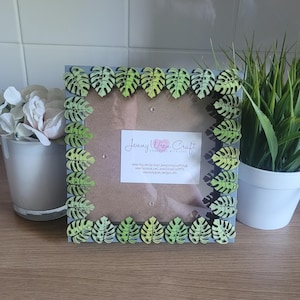 May include: Square grey picture frame decorated with green and yellow leaf cutouts. The frame has a clear glass insert and a small white label with the text "Jenny Wren Craft".