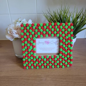 May include: A red and green pom-pom picture frame with a white background. The frame is decorated with red and green pom-poms in a checkered pattern. The frame has a small card inside with the text "Jenny Wren Craft Handmade with Love" and contact information.