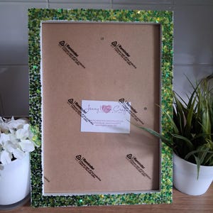 May include: A rectangular picture frame with a green glitter border. The frame has a white interior and a brown backing. A white label with text is visible in the center of the frame. A small plant in a white pot is on the right.