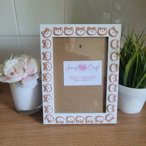 May include: White picture frame with a brown border decorated with cartoon bear faces. The frame has a brown mat and a clear glass insert. The frame is sitting on a wooden surface. The text "Jenny Wren Craft Handmade with Love" is printed on the mat.