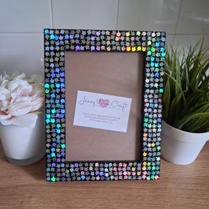 May include: Black rectangular picture frame with iridescent embellishments. The frame has a brown backing and a white card with text. A white pot with flowers and a small potted plant are in the background.
