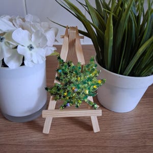 May include: A green, glittery Christmas tree decoration with star-shaped accents, displayed on a small wooden easel. The background includes white flowers in a white pot and a potted green plant.