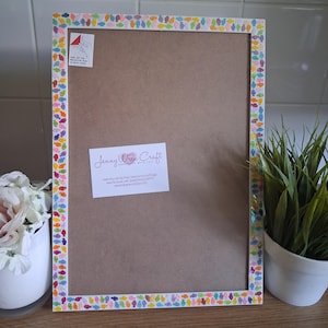 May include: A rectangular picture frame with a white border decorated with a colorful pattern of small fish. The center is a brown board featuring a white sticker with the text Jenny Wren Craft and a small square instruction label in the corner.