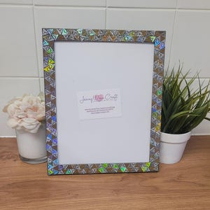 May include: A silver picture frame with a holographic border featuring a repeating pattern of triangles. The frame is decorated with a silver and gold glitter finish. The frame is sitting on a wooden surface.