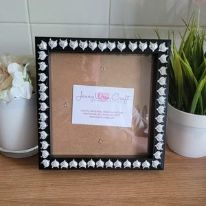 May include: Black square picture frame with silver leaf-shaped detailing around the edge. The frame contains a brown backing and a white card with the text "Jenny Wren Craft" and social media details.