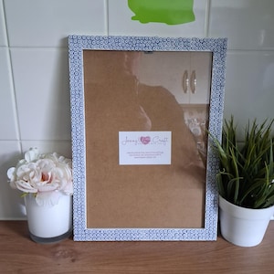 May include: A rectangular picture frame with a blue and white patterned border. The frame has a clear glass front and a brown backing. A white label with the text "Jenny Wren Craft" is affixed to the glass. A small white pot with pink flowers and a small potted plant are next to the frame.