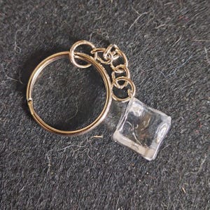 May include: A silver-coloured keyring with a clear, square-shaped charm. The keyring has a split ring and a small chain connecting to the charm. The background is a dark, textured surface.