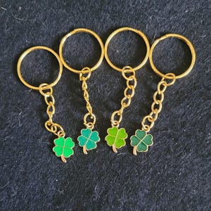 May include: Four gold-coloured keyrings, each with a shamrock charm. Each keyring features a gold ring, a gold chain, and a green shamrock pendant. The shamrocks have varying shades of green against a dark background.